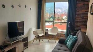 A modern living room with a flat-screen TV, a sofa, two white chairs, a large window with a city view, and a hanging light fixture. - Lovely one bedroom condo in Laureles (Medellín)
