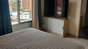 A hotel room with a large bed, a television on a wooden cabinet, and a view of buildings through a window. - Lovely one bedroom condo in Laureles (Medellín)
