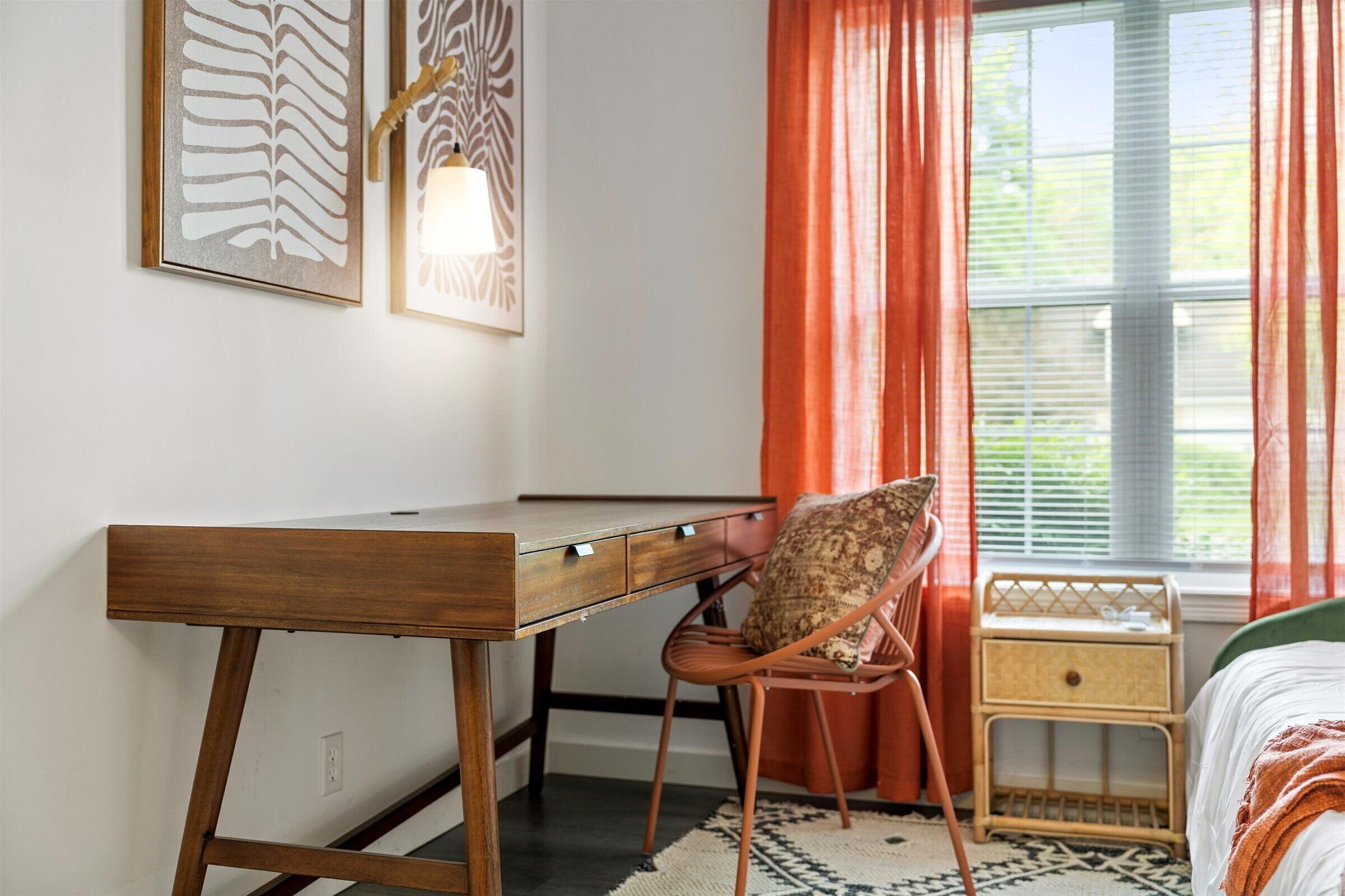 Une chambre à coucher avec un bureau en bois, une chaise en osier, une table de chevet et une fenêtre avec des rideaux.