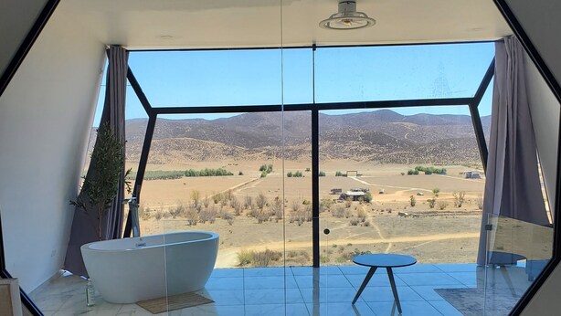 Ein Schlafzimmer mit einem großen Bett, einer Badewanne und Blick auf eine Wüstenlandschaft durch ein sechseckiges Fenster.