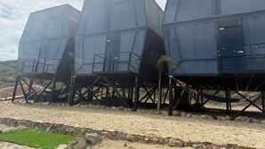 Drei moderne, geometrische Gebäude mit Metallgerüsten und transparenten Flächen, gelegen an einem Sandstrand mit einer Steinbordsteinkante und grünem Rasen davor.