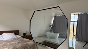 Ein Schlafzimmer mit einem großen, achteckigen Spiegel, einem Bett mit hölzernem Kopfteil, einem Nachttisch und einem Badezimmer mit Badewanne und Dusche.