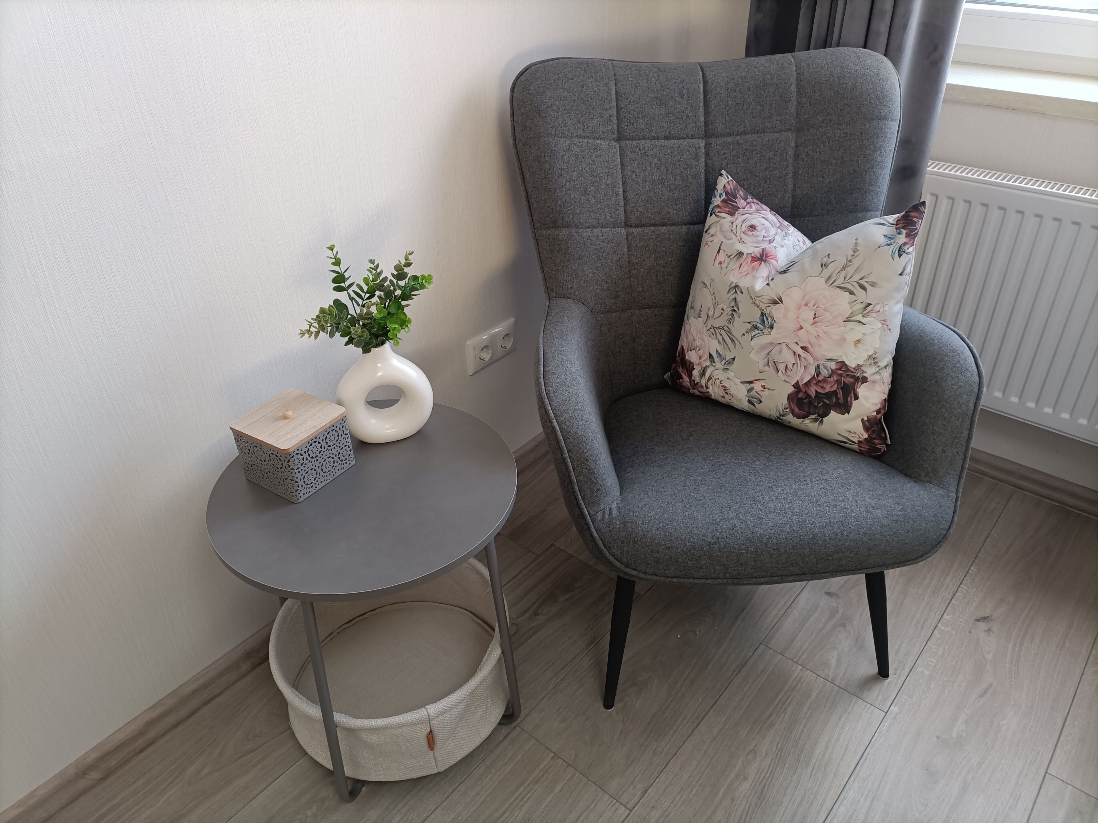 Un fauteuil à revêtement gris agrémenté d’un coussin à motifs floraux, une petite table ronde sur laquelle est posé un vase contenant des plantes vertes, et un panier en osier.