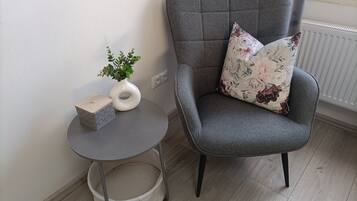 Un fauteuil à revêtement gris agrémenté d’un coussin à motifs floraux, une petite table ronde sur laquelle est posé un vase contenant des plantes vertes, et un panier en osier.