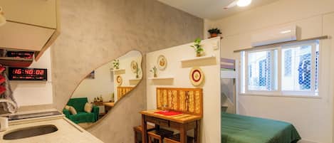 Una habitación compacta con kitchenette, una cama con cubrecama verde, un espejo y un ventilador de techo.