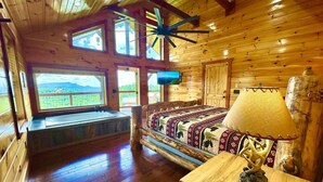 A log cabin bedroom with a large bed, a wooden desk, a television, and a view of the outdoors. - New Amazing View Cabin, 5 miles to Dollywood, 12 miles to Smokies, Sleeps 11 (Sevierville)