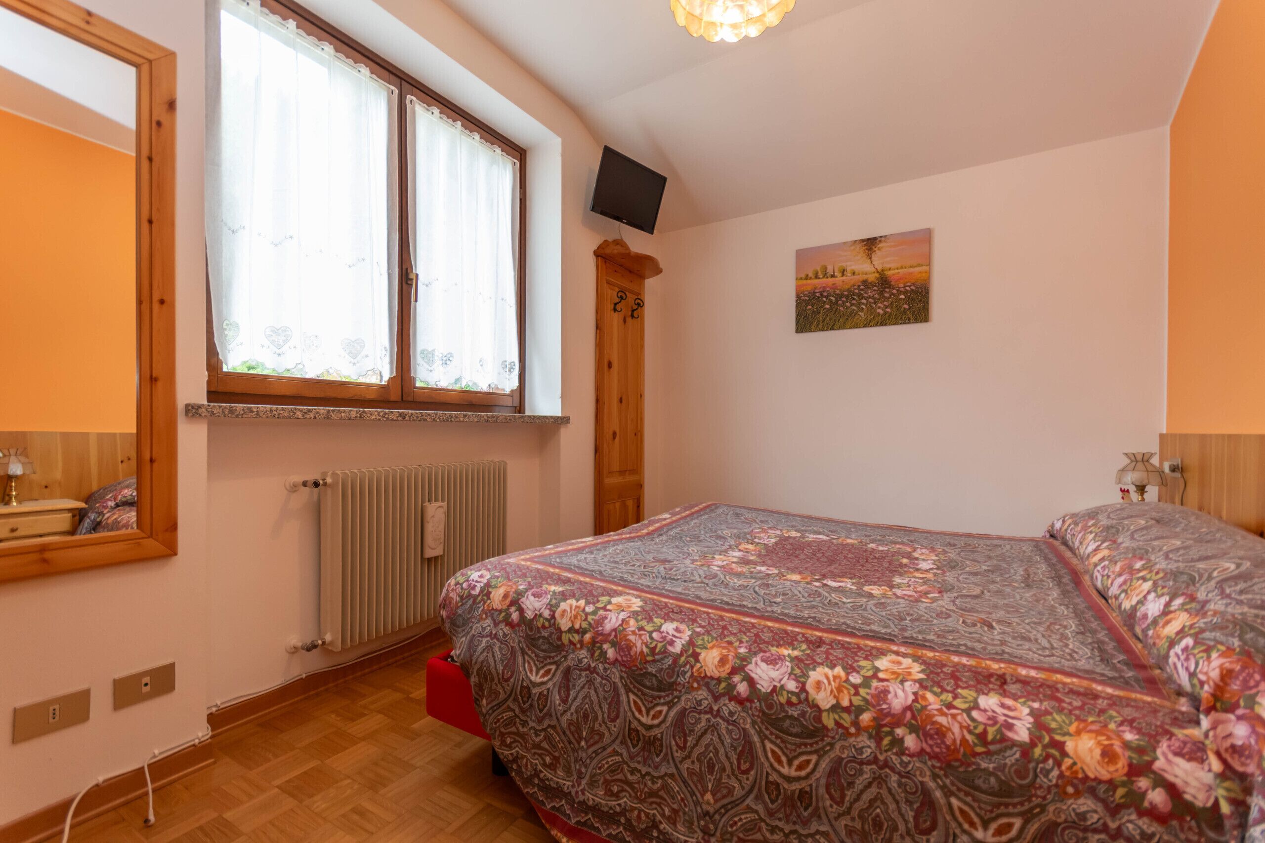 A bedroom with a large bed, a window with curtains, a radiator, a wooden wardrobe, a painting on the wall, and a television mounted on the wall.