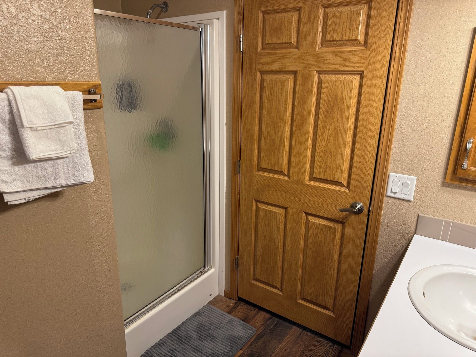 Un baño con puerta de madera, cabina de ducha de vidrio, lavamanos blanco y toallero con toallas dobladas.