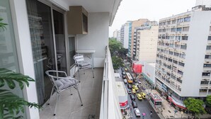 Un balcón con dos sillas, una mesa y vistas a una calle concurrida con edificios y vehículos.