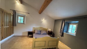 Un dormitorio con cama, mesita de noche, armario y dos ventanas.