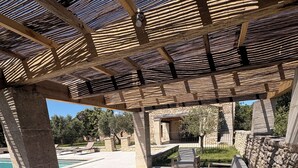 Un espacio de estar al aire libre cubierto, con pilares de piedra, una piscina al fondo y un muro de piedra.