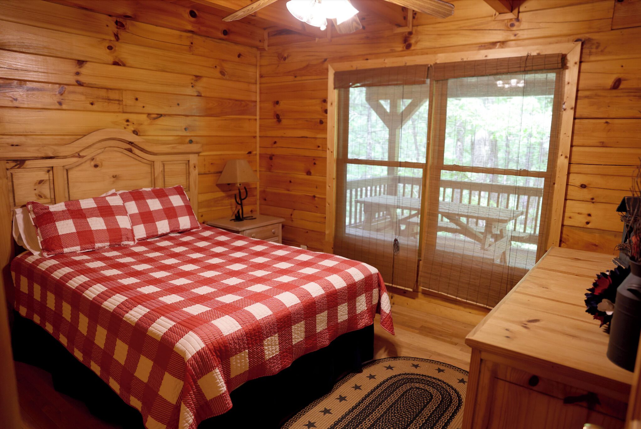 A wooden cabin bedroom with a checkered bedspread, a nightstand with a lamp, and a view of an outdoor deck through a screened door.