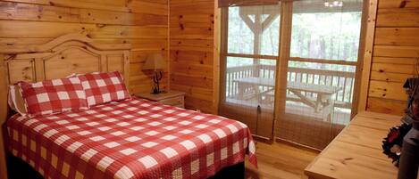 A wooden cabin bedroom with a checkered bedspread, a nightstand with a lamp, and a view of an outdoor deck through a screened door.