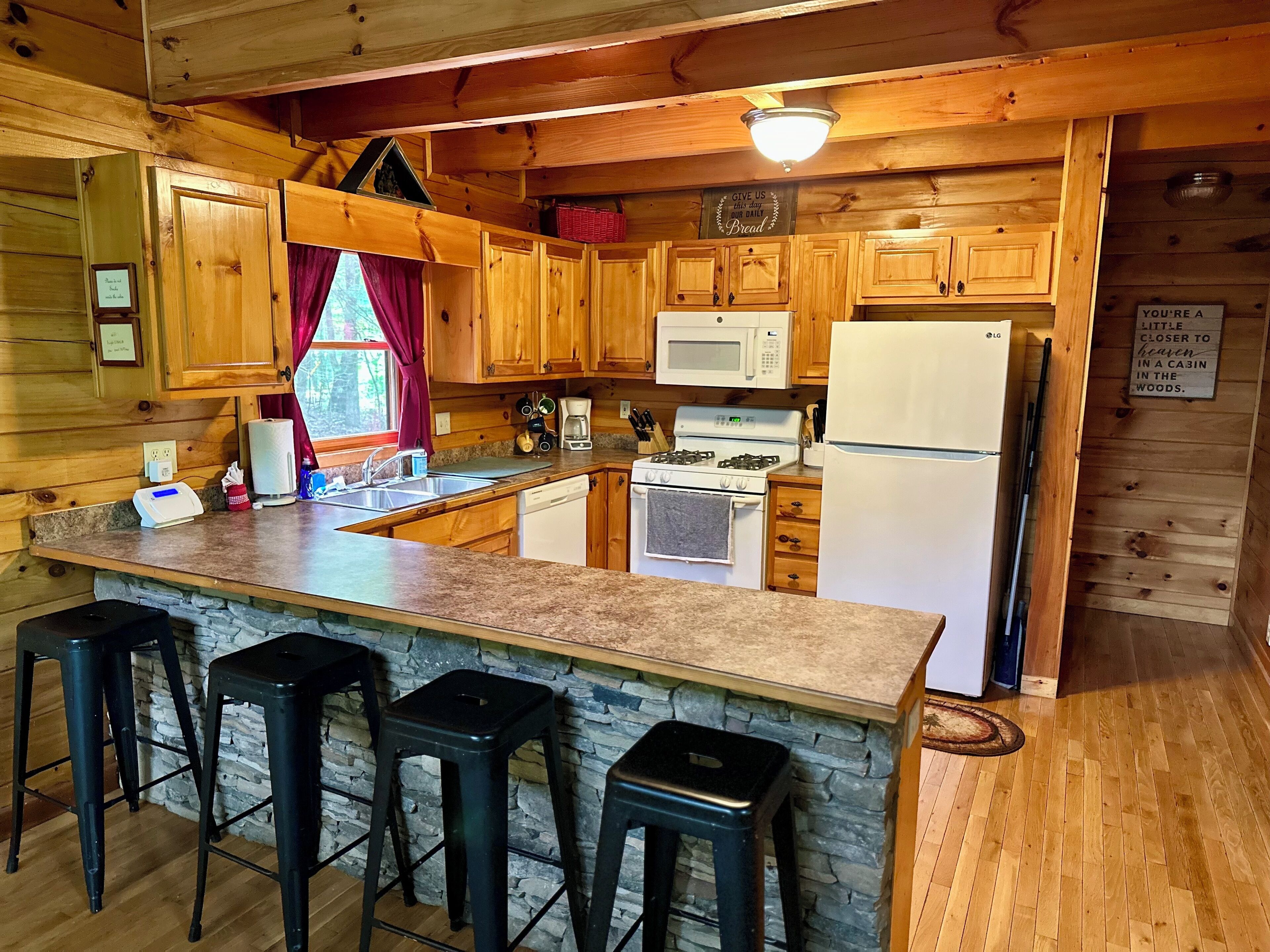 A cabin-style kitchen with wooden cabinets, a granite countertop, a gas stove, a microwave, and a refrigerator. There are three bar stools and a stone wall.