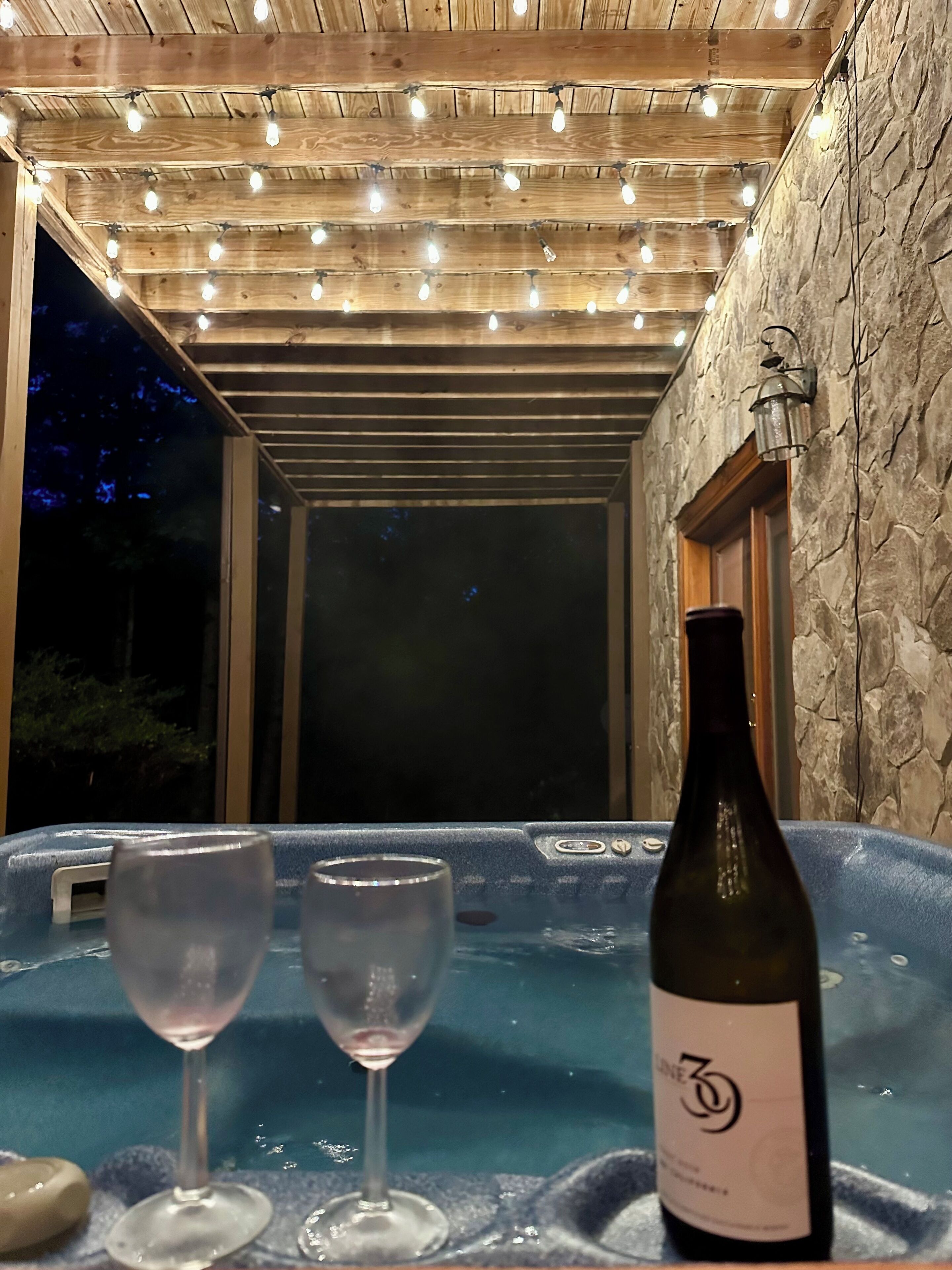A hot tub with two glasses and a bottle of wine, wooden ceiling, stone wall, and a lantern.