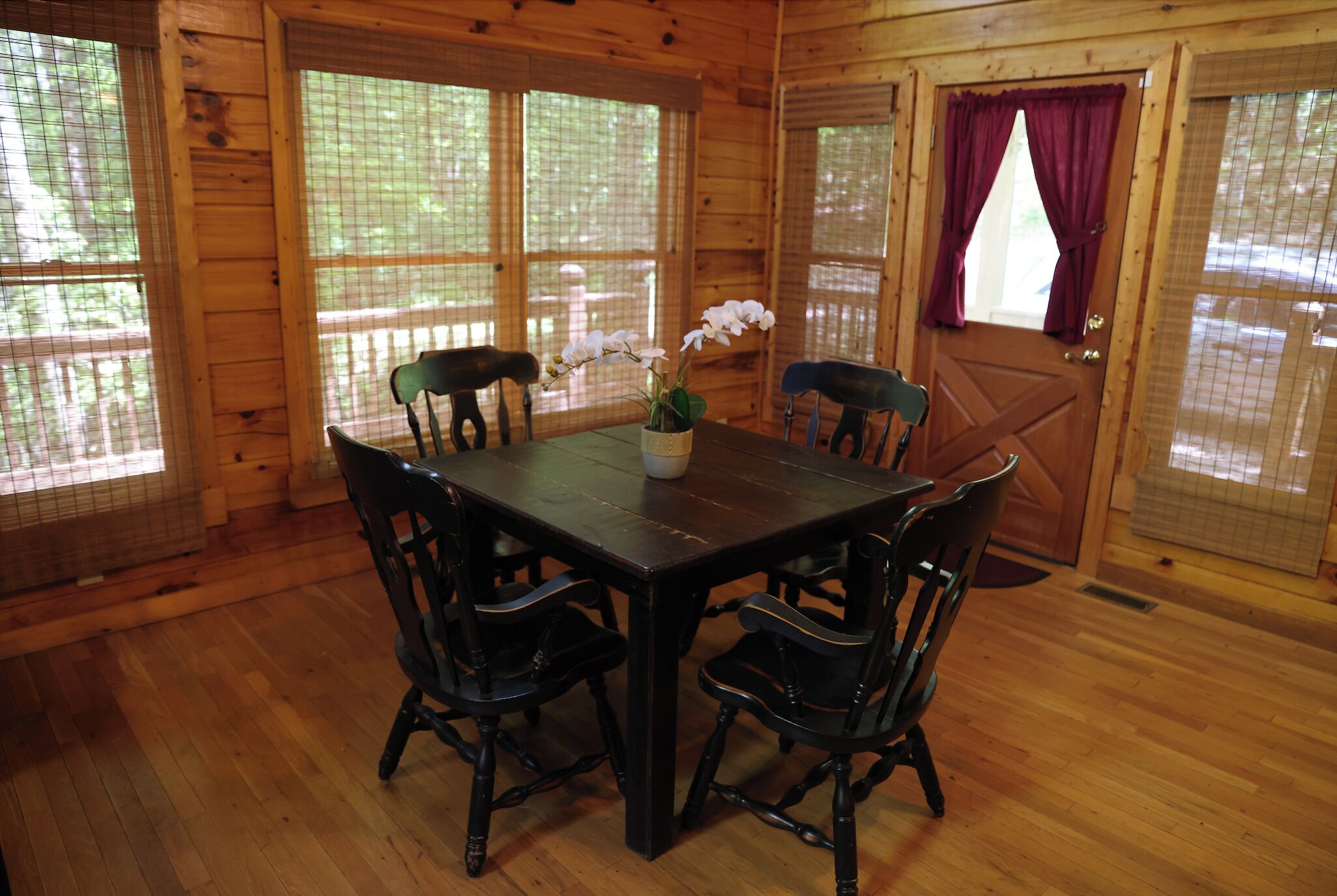 A wooden dining room with a table and chairs, large windows with curtains, and a door with a screen.