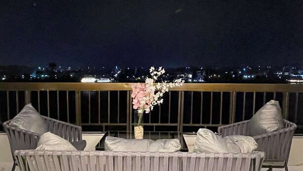 A balcony with two grey wicker chairs, a small table with a vase of flowers, and a view of a cityscape at night.