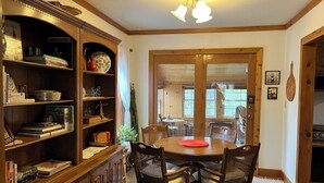A dining area with a wooden table and chairs, a bookshelf with various items, and a window with a view of the outdoors.