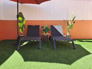 Two lounge chairs, a small table with a drink, and potted plants in an outdoor seating area with artificial grass and a corrugated metal wall.