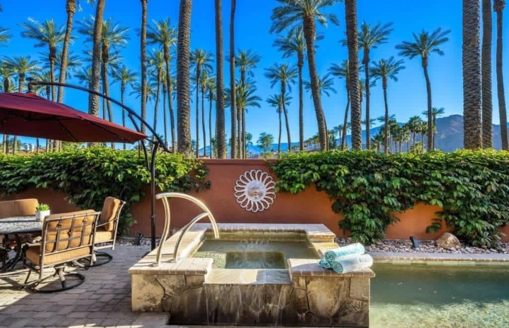 Un área de piscina con fuente de agua, rodeada de palmeras y un patio con mobiliario exterior.