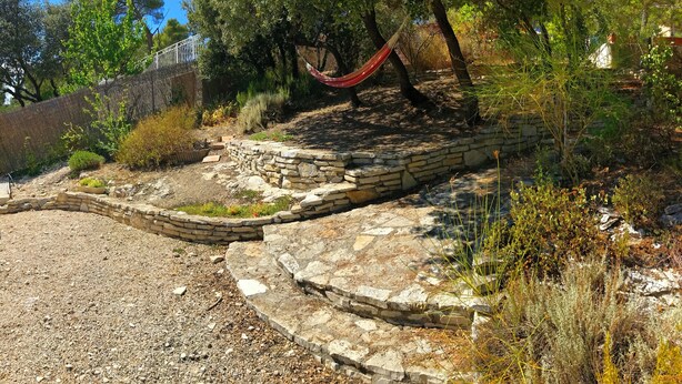 Un muro de piedra con una hamaca, rodeado de vegetación seca y árboles.