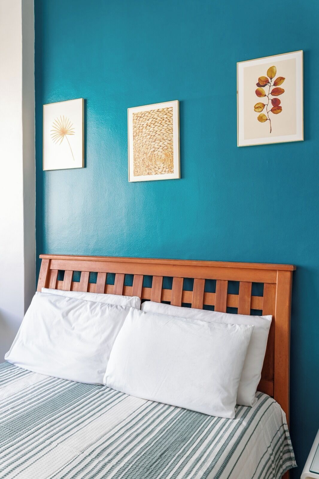 Un lit avec des oreillers blancs et une couvre-lit à rayures, une tête de lit en bois et trois œuvres d’art encadrées sur un mur de couleur turquoise.