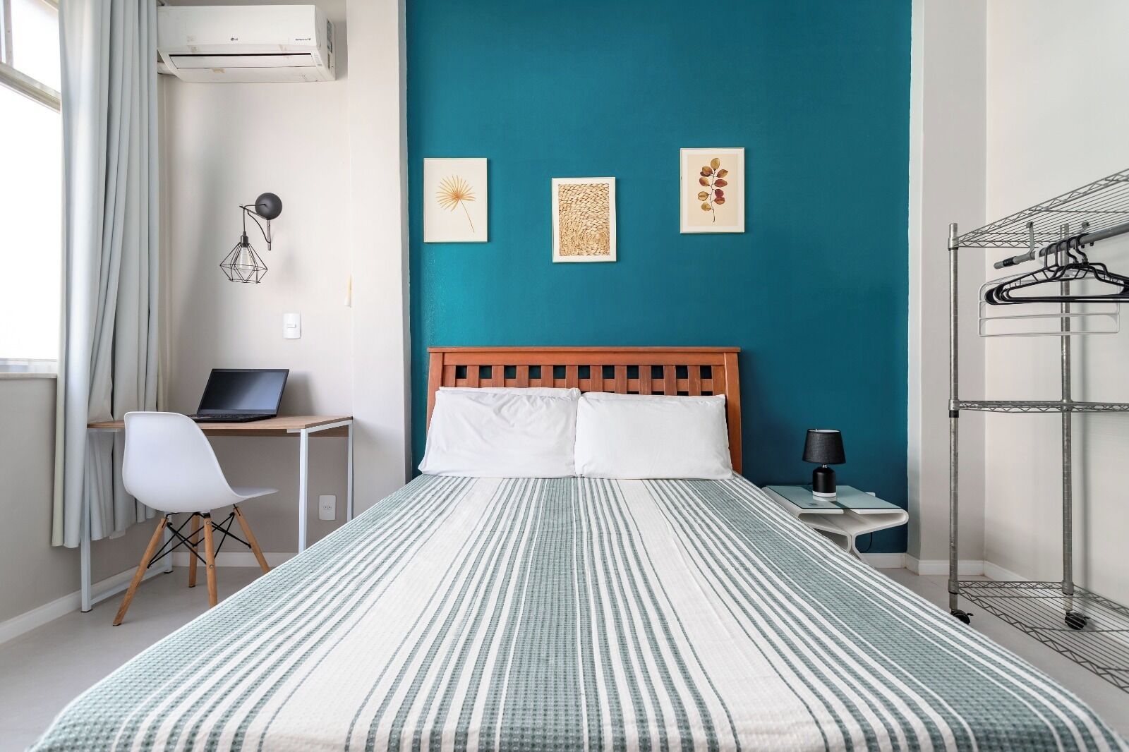 A bedroom with a bed, a desk with a laptop, a chair, a nightstand with a lamp, and a metal rack.