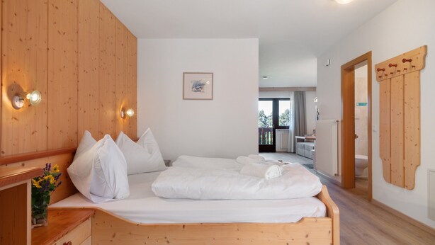 Una cama de madera con ropa de cama blanca, una mesita de noche con un florero y un armario de madera con un espejo.