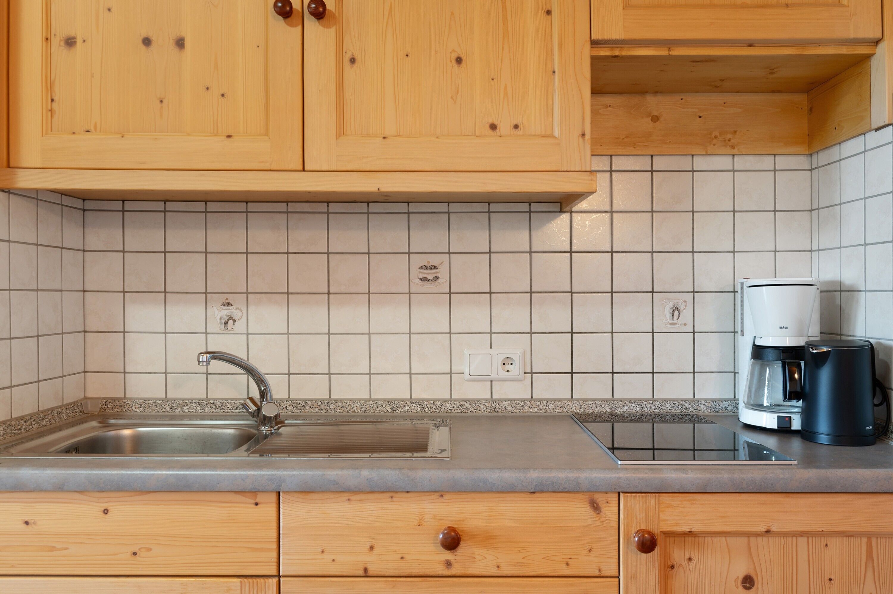 Cozinha com armários de madeira, pia de aço inoxidável, cafeteira e painel de azulejos.
