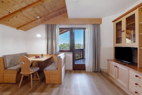 A cozy room with a wooden ceiling, a dining table with chairs, a sofa, a television on a wooden cabinet, and a window with a view.