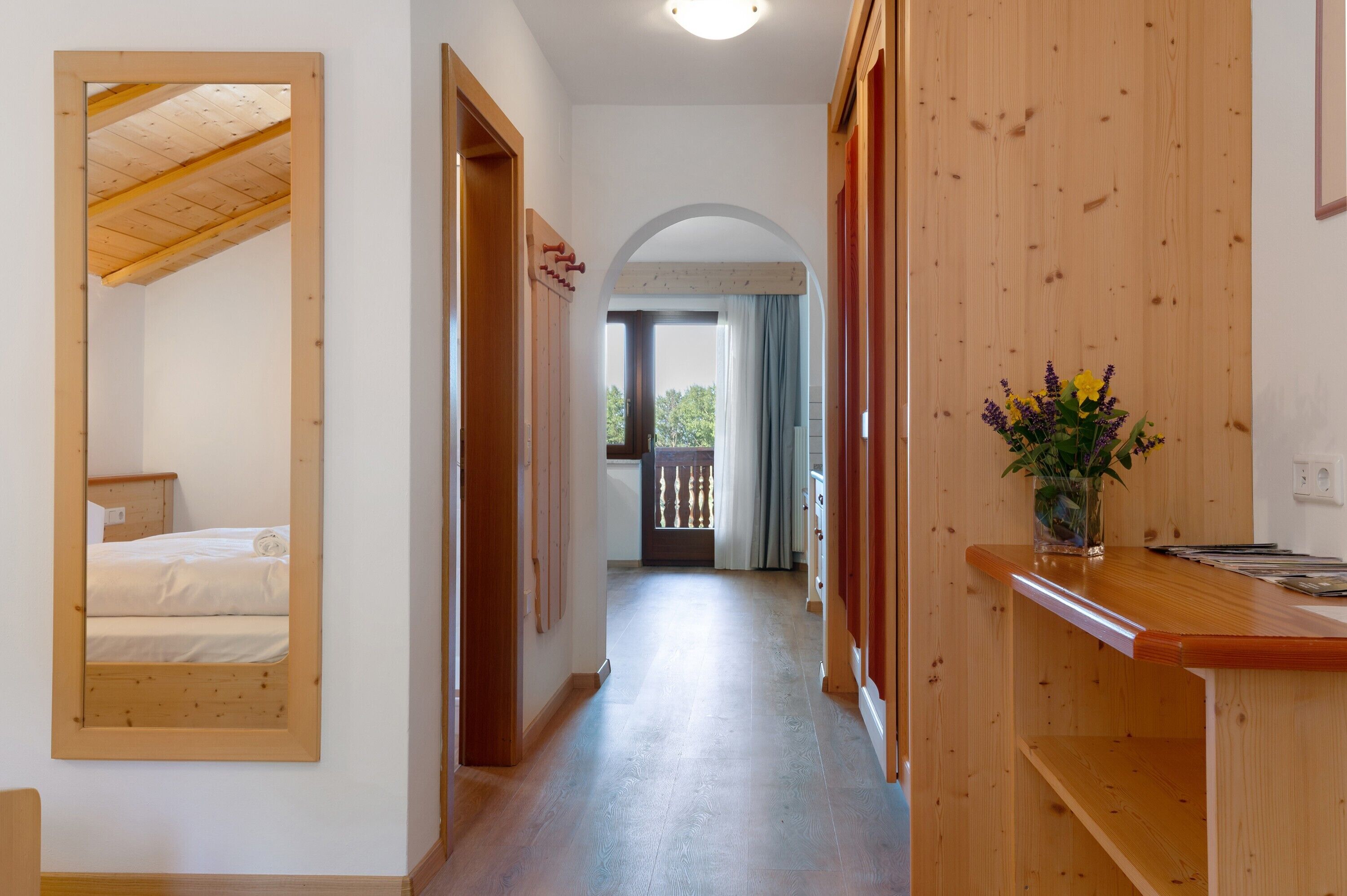 A hallway with a wooden floor, a bed in the left room, and a wooden counter with a vase of flowers.
