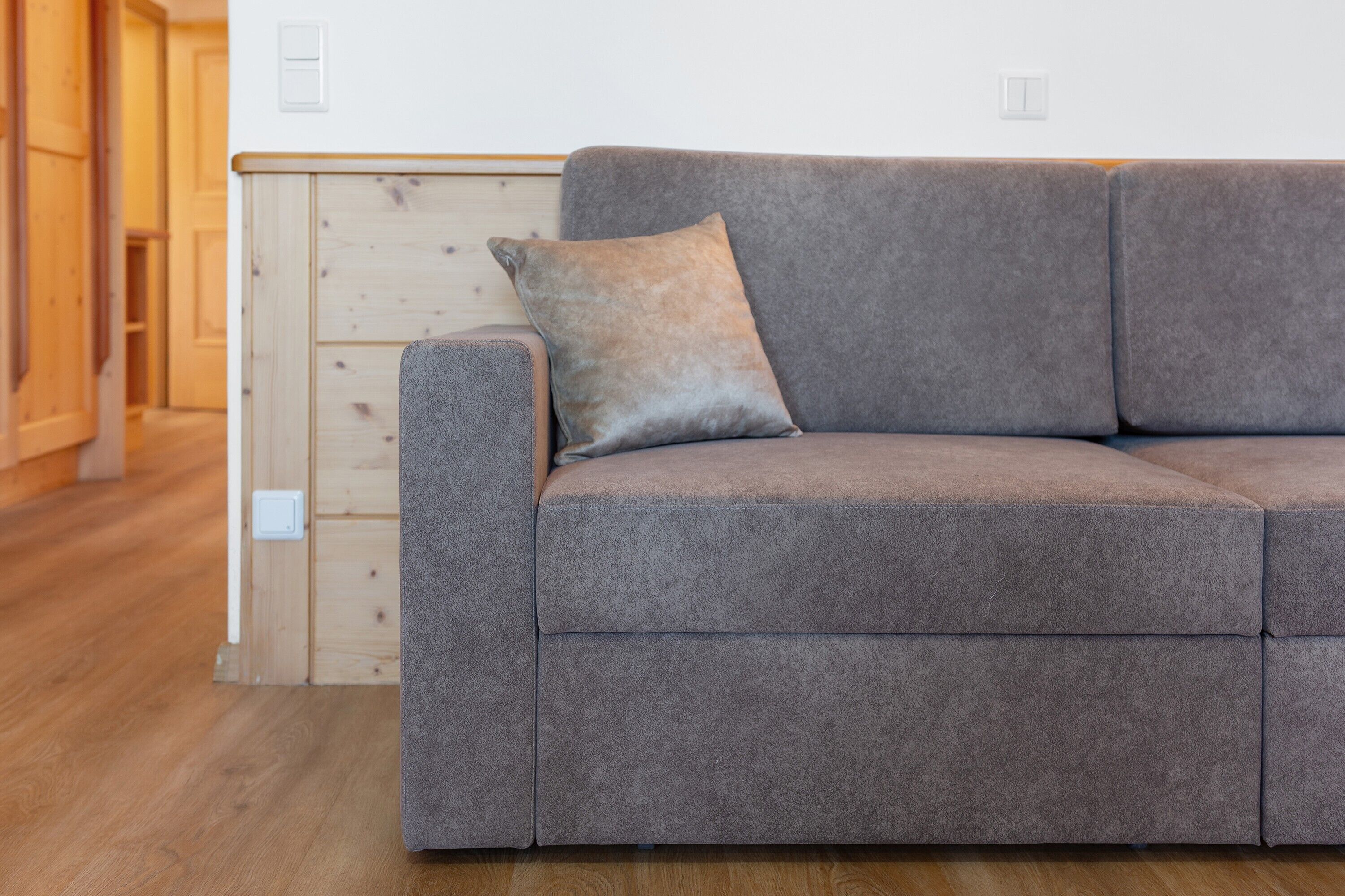A grey sofa with a single cushion, a wooden floor, and a white wall with a light switch.