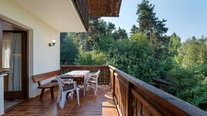 Un balcón con barandilla de madera, una mesa con sillas y vistas a una zona boscosa.