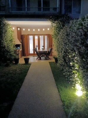 A pathway leading to a dining area with a table and chairs, surrounded by greenery and a building entrance.