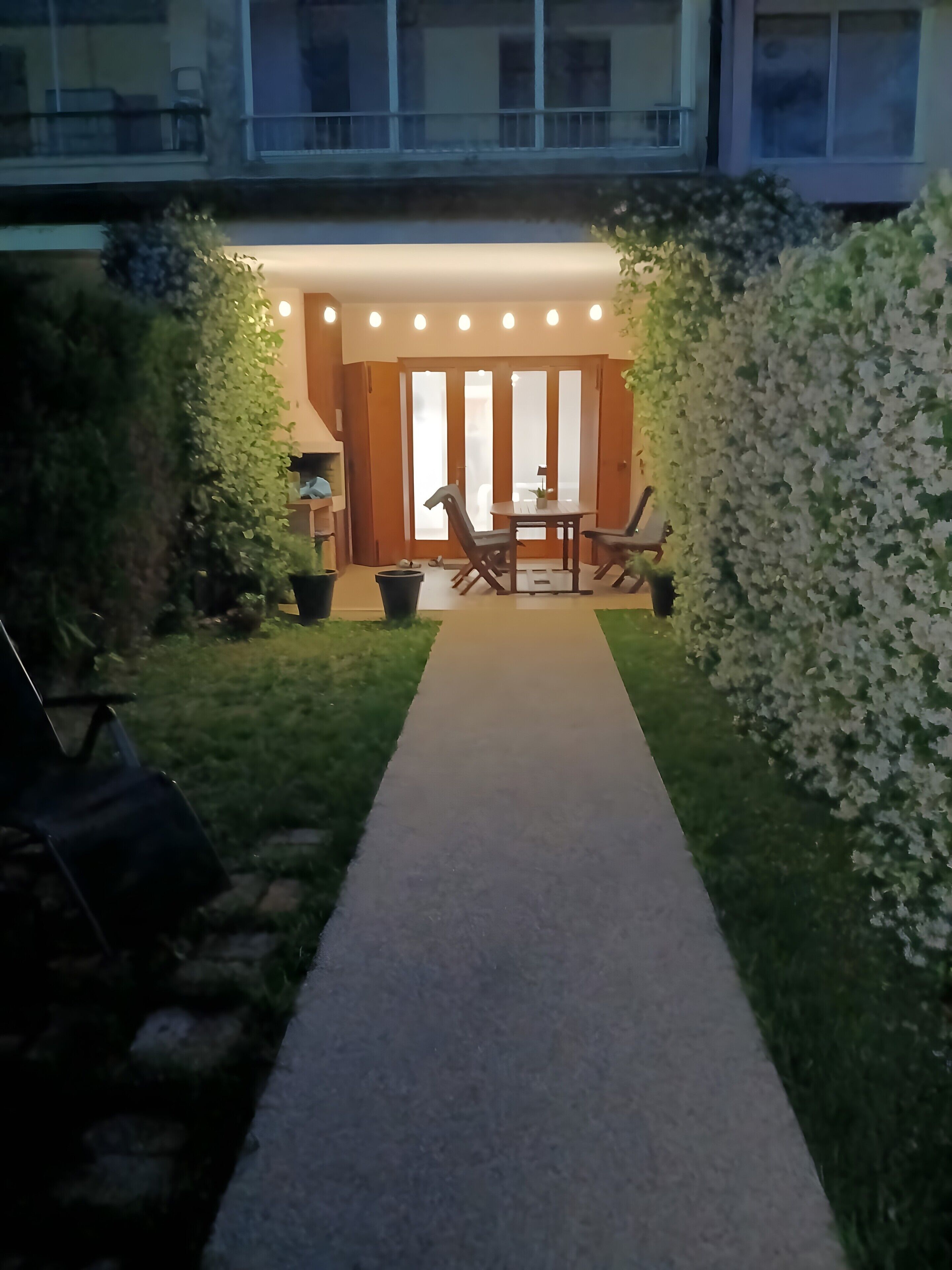 Un sendero lleva a una mesa y sillas de madera, rodeado de vegetación y la entrada de un edificio.