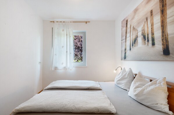 Un dormitorio con cama, ventana con cortinas, un cuadro en la pared y una lámpara de noche.
