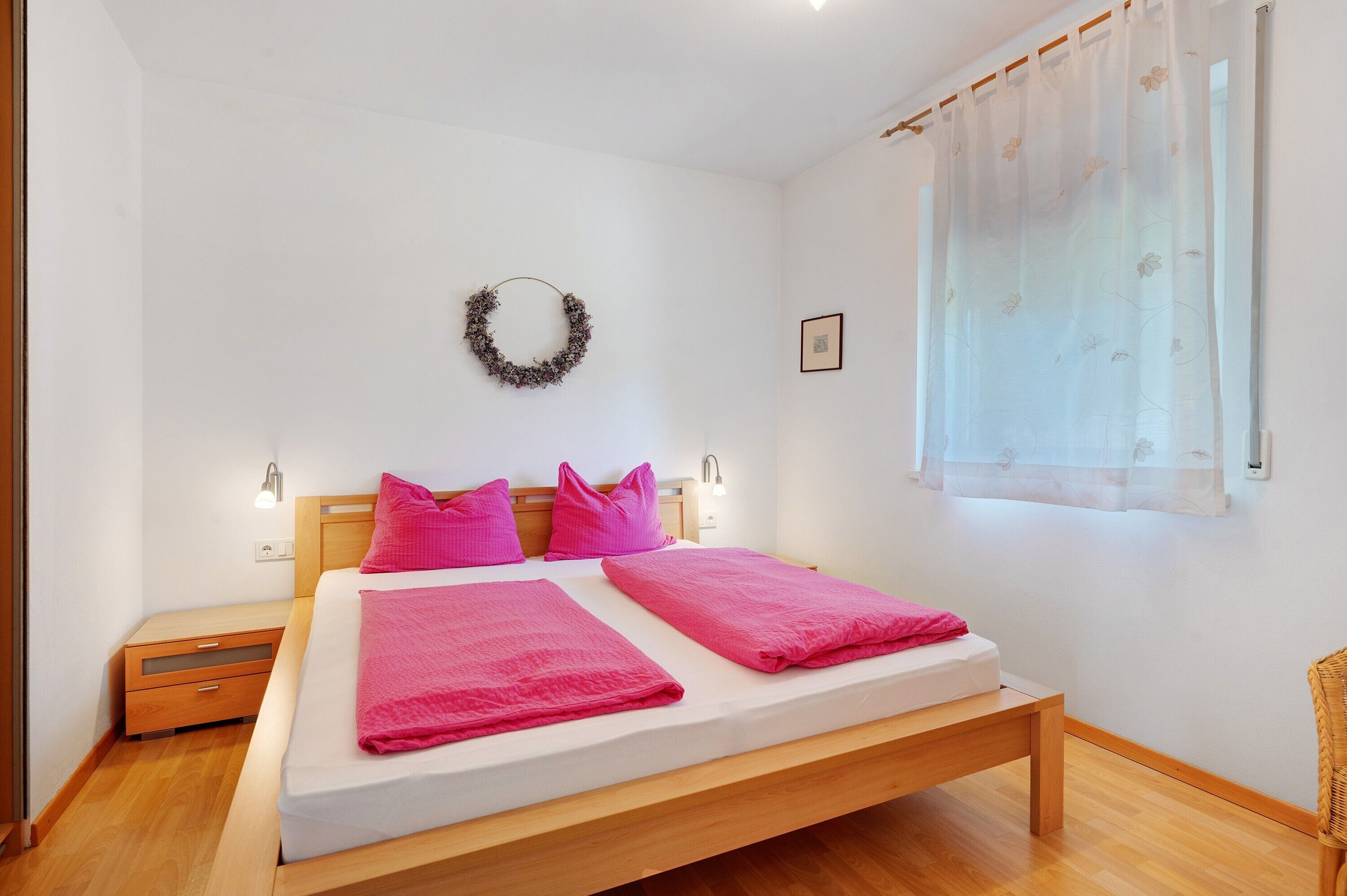 Une chambre avec un lit simple, une tête de lit en bois, deux oreillers roses, un matelas blanc, une table de chevet en bois, un petit tiroir, une lampe fixée au mur, un tableau encadré, une fenêtre avec des rideaux légers et une guirlande décorative accrochée au mur.