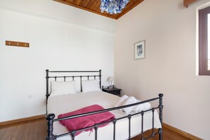 A bedroom with a bed, a nightstand, a lamp, a red blanket, a wooden floor, a white wall, a ceiling with a wooden beam, and a framed picture.