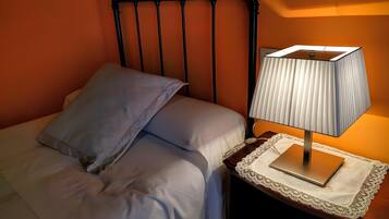 Una cama con ropa blanca y una sola almohada azul, una mesita de noche con lámpara y un cabecero de madera.
