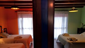 Two twin beds with towels, a nightstand, and a lamp in a room with wooden beams and a window with sheer curtains.