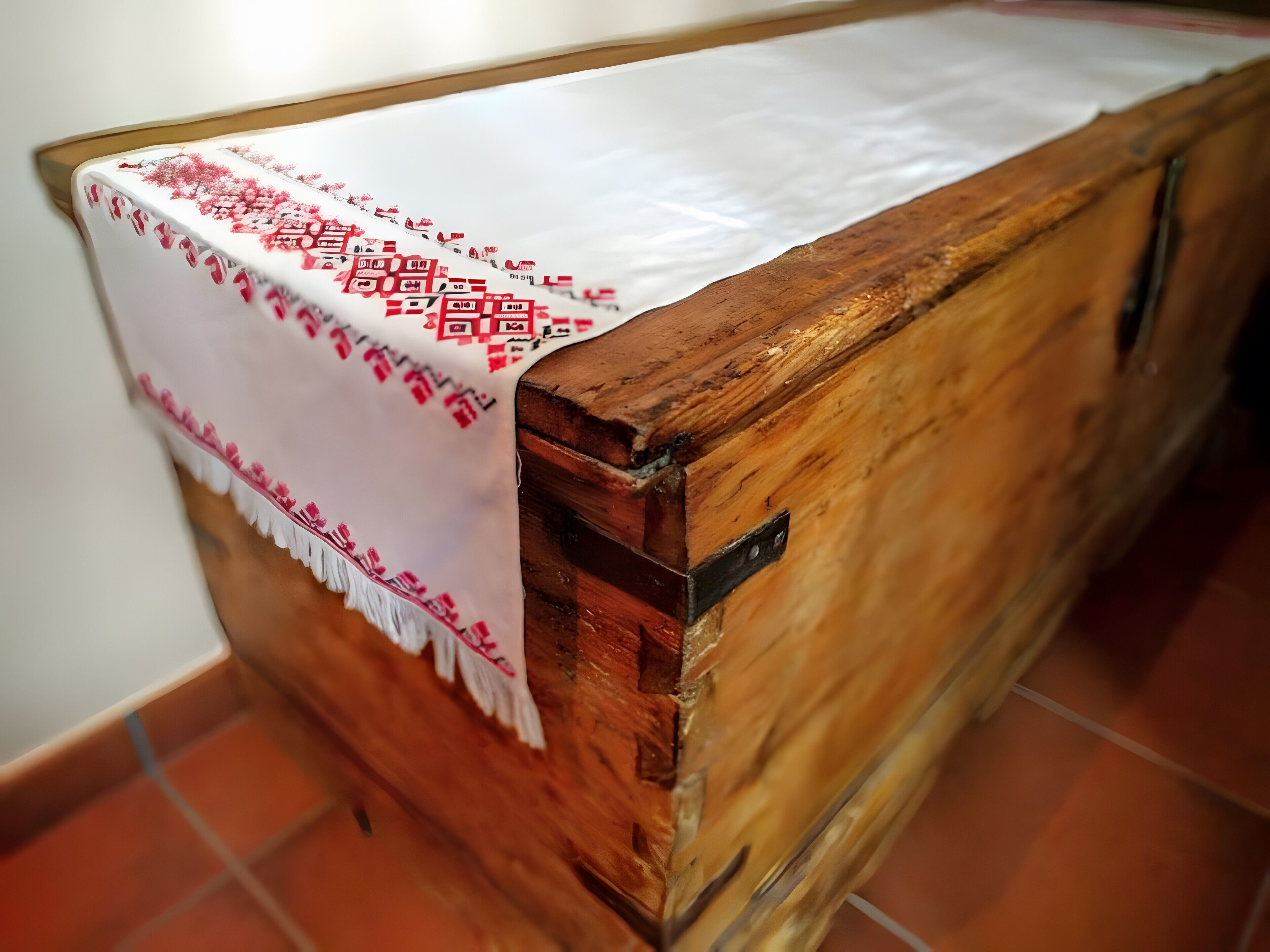 Une boîte en bois recouverte d’un tissu blanc orné de motifs et de lettres rouges, posée sur un sol carrelé.