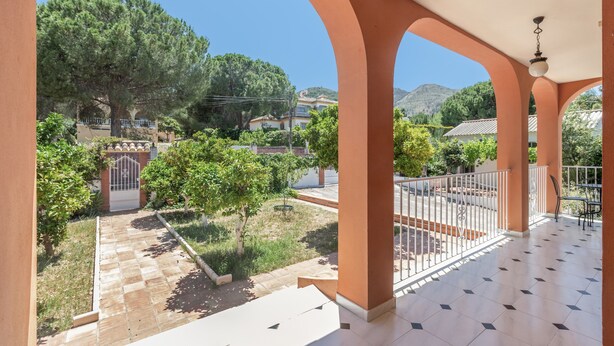 Ein überdachter Außenbereich mit Fliesenboden, Blick auf einen Garten mit Bäumen und einem Tor, sowie ein Berg im Hintergrund.