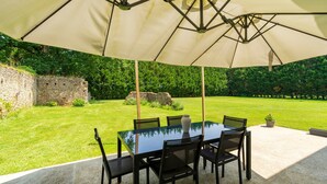 Eine Terrasse im Freien mit Tisch und Stühlen unter einem großen Sonnenschirm, umgeben von einem gepflegten Rasen und einer Steinsmauer.