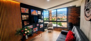 A modern living room with a large window, a black leather sofa, a wooden slatted wall, and various framed artworks. - Stylish Loft in Laureles Gorgeous views  (Medellín)