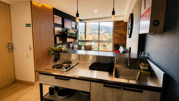 Uma cozinha moderna com bancada de aço inoxidável, fogão embutido e pia. Há uma janela com vista para a paisagem urbana.