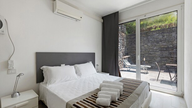 A modern bedroom with a large bed, a bedside table with a lamp, a window with a view of a stone wall and outdoor seating. - Apartment 'Green House' with Private Terrace, Wi-Fi and Air Conditioning (Sestri Levante)