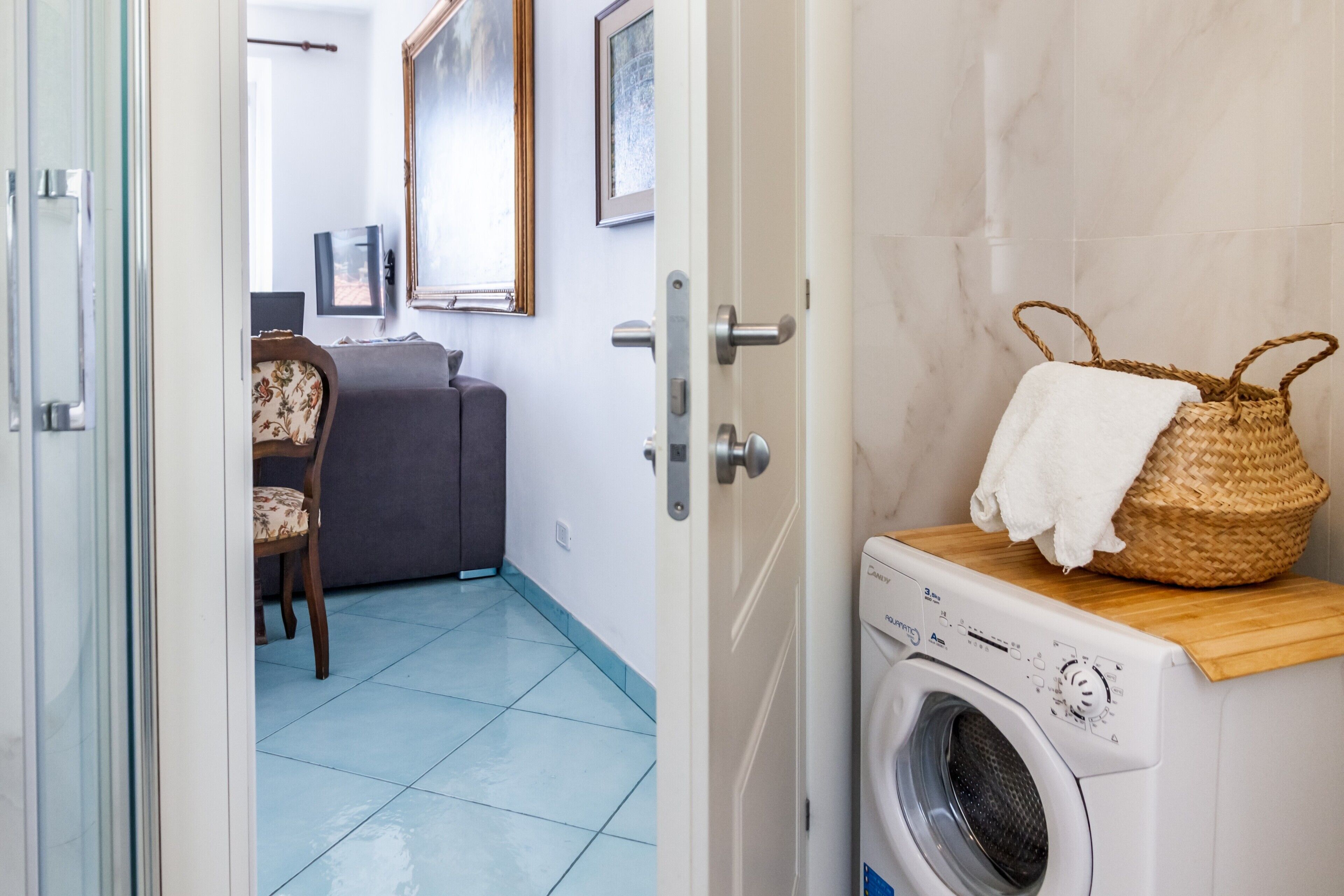 A laundry area with a washing machine, a wicker basket, and a grey sofa in the background.