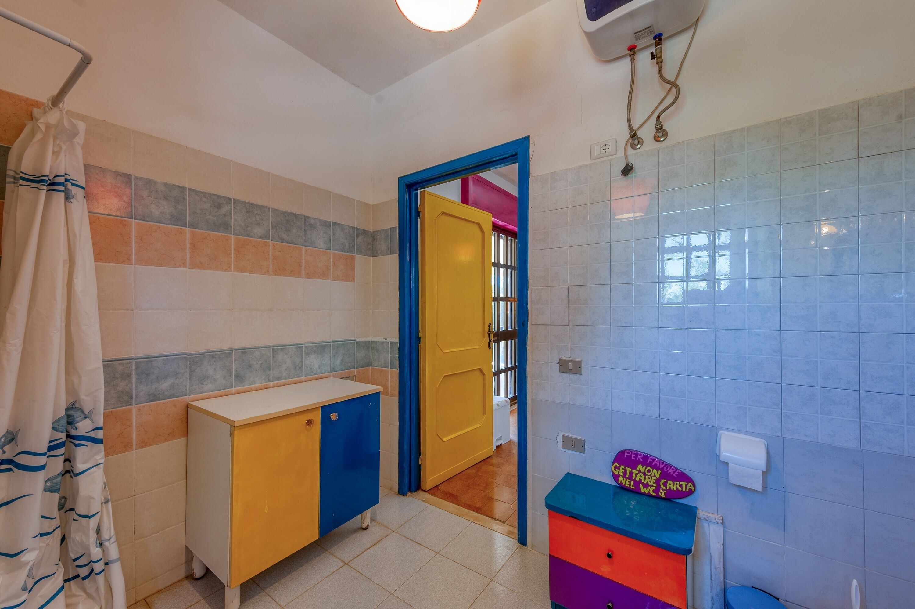 A bathroom with a tiled wall, a yellow door, a blue-framed door, a white sink, and a colorful cabinet.