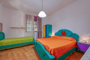 A bedroom with a bed, a bedside table, a nightstand, a window with curtains, and a wall-mounted shelf.