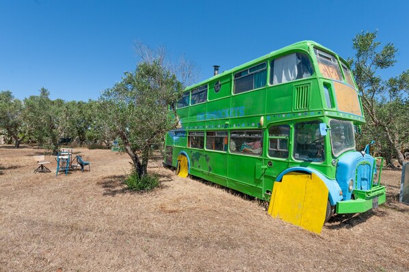 A green double-decker bus parked in a dry, grassy area with trees and a clear blue sky.
